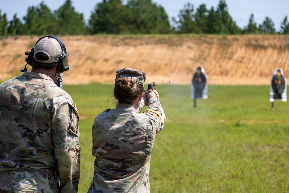 Two military personnel in OCP (Operational Camouflage Pattern) uniforms are on an outdoor shooting range. A female soldier is actively firing a pistol at humanoid silhouette targets downrange, with a spent casing visibly ejecting from the firearm. A male soldier wearing hearing protection and a gray cap stands beside her, observing or supervising.