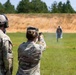 Two military personnel in OCP (Operational Camouflage Pattern) uniforms are on an outdoor shooting range. A female soldier is actively firing a pistol at humanoid silhouette targets downrange, with a spent casing visibly ejecting from the firearm. A male soldier wearing hearing protection and a gray cap stands beside her, observing or supervising.
