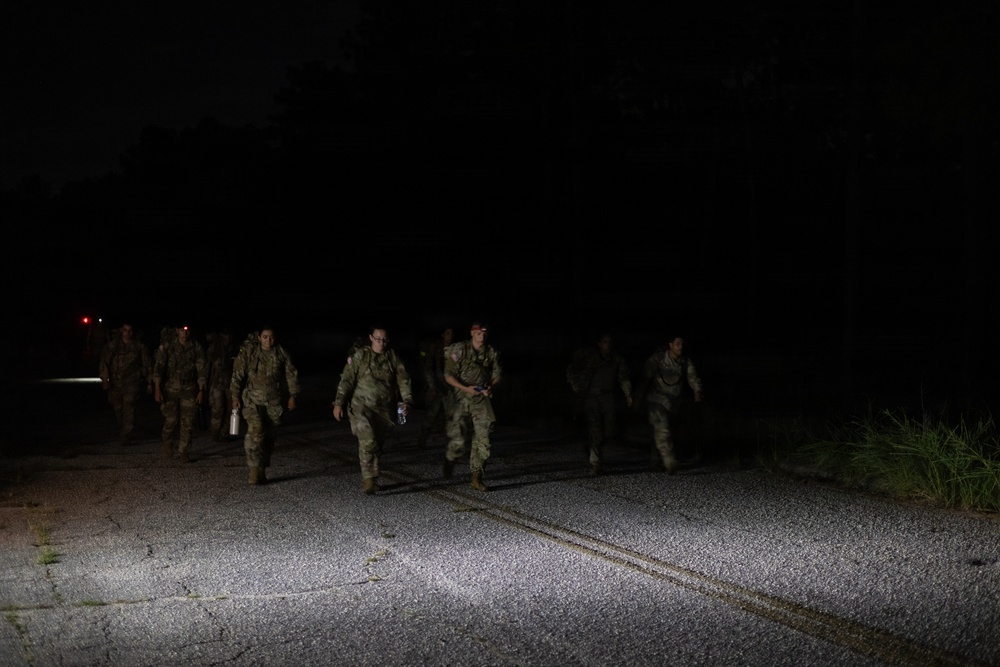 A group of military personnel in camouflage uniforms are marching along a paved road at night. They appear to be conducting a nighttime road march or ruck march exercise, illuminated by a light source from behind the camera. The soldiers are carrying packs and equipment consistent with a training exercise.
