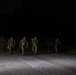 A group of military personnel in camouflage uniforms are marching along a paved road at night. They appear to be conducting a nighttime road march or ruck march exercise, illuminated by a light source from behind the camera. The soldiers are carrying packs and equipment consistent with a training exercise.