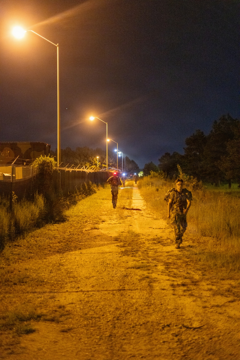 A nighttime scene showing military personnel in camouflage uniforms conducting what appears to be a ruck march or training exercise along a dirt path. The path is illuminated by a series of orange sodium-vapor street lamps stretching into the distance. At least two soldiers are visible in the foreground, with additional personnel visible further down the path.