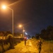A nighttime scene showing military personnel in camouflage uniforms conducting what appears to be a ruck march or training exercise along a dirt path. The path is illuminated by a series of orange sodium-vapor street lamps stretching into the distance. At least two soldiers are visible in the foreground, with additional personnel visible further down the path.