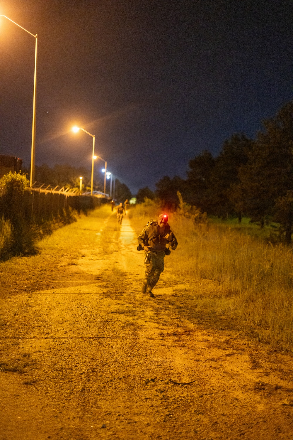 A nighttime outdoor scene showing at least two individuals in military-style gear running or marching along a dirt/gravel path illuminated by overhead street lamps. The foreground figure wears a red light on their helmet and carries equipment consistent with military training or operations. The setting appears to be a military installation or training area, bordered by a fence line on the left and vegetation on the right.