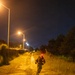 A nighttime outdoor scene showing at least two individuals in military-style gear running or marching along a dirt/gravel path illuminated by overhead street lamps. The foreground figure wears a red light on their helmet and carries equipment consistent with military training or operations. The setting appears to be a military installation or training area, bordered by a fence line on the left and vegetation on the right.