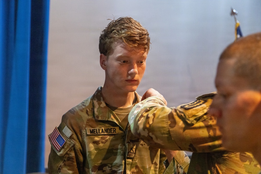 A U.S. Army soldier named Mellander is visible in the foreground wearing OCP (Operational Camouflage Pattern) uniform with a U.S. flag patch on the left shoulder. A second soldier, partially visible in the foreground right, appears to be pinning or adjusting something on Mellander's uniform, suggesting a military ceremony or promotion event. The setting appears to be an indoor venue with a blue curtain and what appears to be a military flag in the background.
