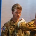 A U.S. Army soldier named Mellander is visible in the foreground wearing OCP (Operational Camouflage Pattern) uniform with a U.S. flag patch on the left shoulder. A second soldier, partially visible in the foreground right, appears to be pinning or adjusting something on Mellander's uniform, suggesting a military ceremony or promotion event. The setting appears to be an indoor venue with a blue curtain and what appears to be a military flag in the background.