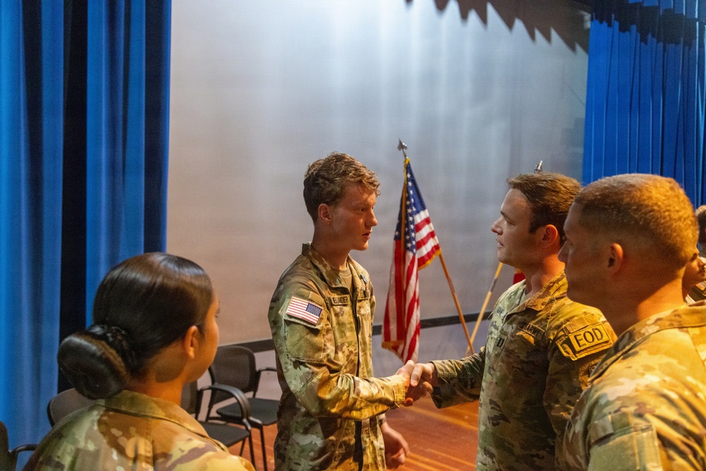A group of U.S. Army soldiers in Army Combat Uniforms (ACU/OCP pattern) are gathered in what appears to be an auditorium or ceremony hall. Two soldiers are shaking hands in the foreground, suggesting a formal or ceremonial interaction. An American flag and what appears to be a unit flag are visible on a stage in the background.