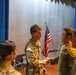 A group of U.S. Army soldiers in Army Combat Uniforms (ACU/OCP pattern) are gathered in what appears to be an auditorium or ceremony hall. Two soldiers are shaking hands in the foreground, suggesting a formal or ceremonial interaction. An American flag and what appears to be a unit flag are visible on a stage in the background.