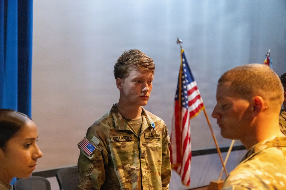 A U.S. Army soldier named Mellander stands at attention during what appears to be a military ceremony or formal event. He is wearing an OCP (Operational Camouflage Pattern) uniform with a medal pinned to his chest, suggesting a decoration or award ceremony. An American flag is visible in the background alongside other personnel in uniform.