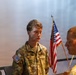 A U.S. Army soldier named Mellander stands at attention during what appears to be a military ceremony or formal event. He is wearing an OCP (Operational Camouflage Pattern) uniform with a medal pinned to his chest, suggesting a decoration or award ceremony. An American flag is visible in the background alongside other personnel in uniform.