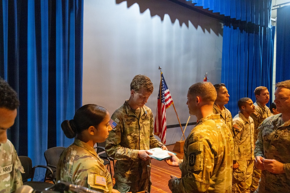 A military ceremony is taking place in an auditorium or assembly hall with blue curtains and a stage backdrop. A soldier identified by nametape as 'MELLANDER' is handing a document or certificate to another soldier in a line, suggesting an awards or recognition ceremony. Multiple U.S. Army personnel in OCP (Operational Camouflage Pattern) uniforms are present, with an American flag visible in the background.