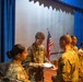 A military ceremony is taking place in an auditorium or assembly hall with blue curtains and a stage backdrop. A soldier identified by nametape as 'MELLANDER' is handing a document or certificate to another soldier in a line, suggesting an awards or recognition ceremony. Multiple U.S. Army personnel in OCP (Operational Camouflage Pattern) uniforms are present, with an American flag visible in the background.
