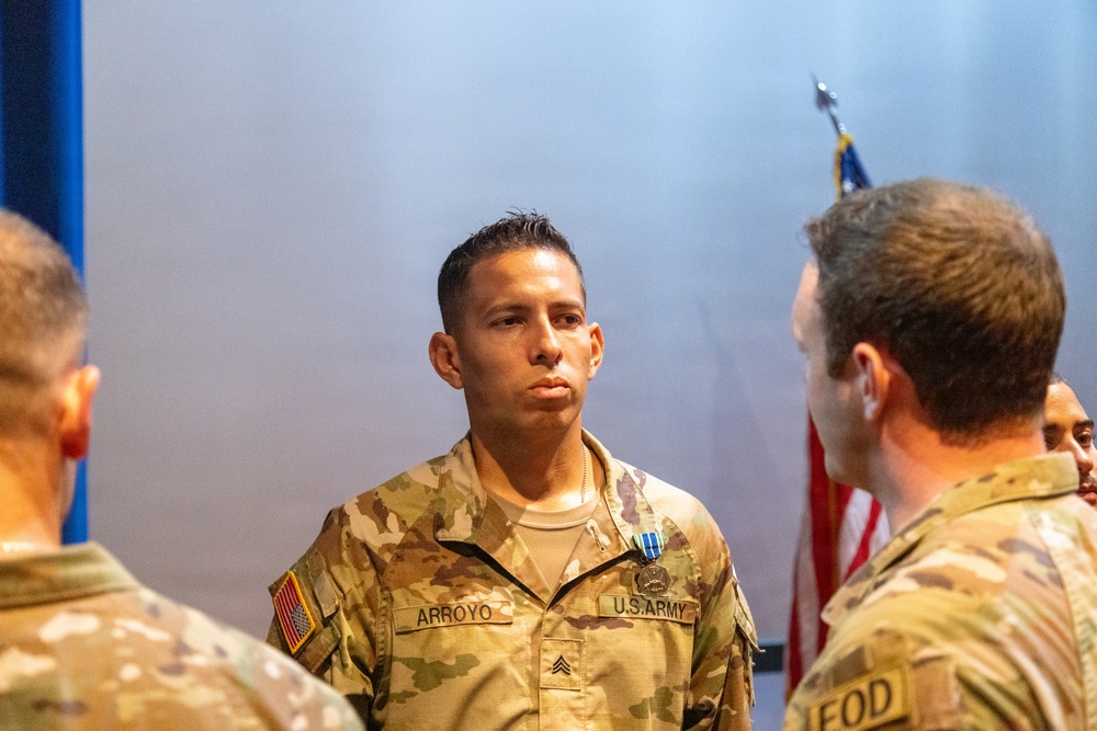 A U.S. Army Sergeant named Arroyo stands at attention during what appears to be a military awards ceremony. He is wearing an Army Combat Uniform (ACU) in OCP (Operational Camouflage Pattern) and has a medal pinned to his chest. Several other soldiers are present, and an American flag is visible in the background.