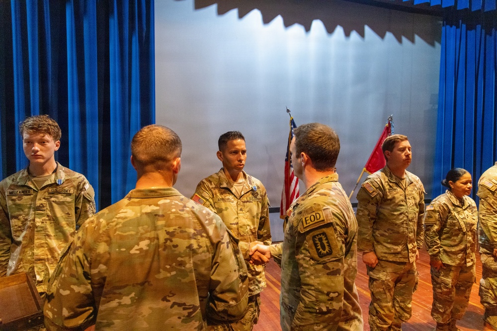 A military ceremony is taking place on a stage or auditorium, where a senior soldier wearing an EOD patch is shaking hands with another soldier named ARROYO, likely presenting an award or commendation. Multiple soldiers in U.S. Army OCP (Operational Camouflage Pattern) uniforms are standing in formation. An American flag and what appears to be a unit flag are visible in the background.