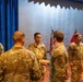 A military ceremony is taking place on a stage or auditorium, where a senior soldier wearing an EOD patch is shaking hands with another soldier named ARROYO, likely presenting an award or commendation. Multiple soldiers in U.S. Army OCP (Operational Camouflage Pattern) uniforms are standing in formation. An American flag and what appears to be a unit flag are visible in the background.