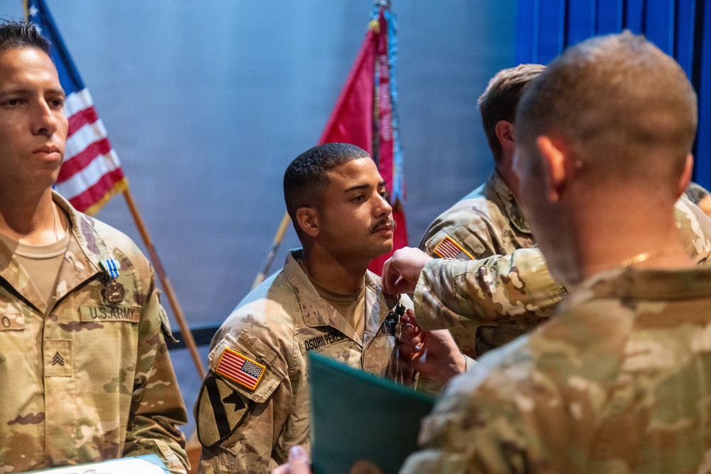 A U.S. Army military ceremony is taking place in which a soldier is receiving a medal or award being pinned onto their uniform by a higher-ranking officer. At least three soldiers in Army Combat Uniforms (ACU/OCP pattern) are visible, with U.S. and unit flags displayed in the background. The setting appears to be an indoor auditorium or ceremony hall.