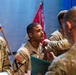 A U.S. Army military ceremony is taking place in which a soldier is receiving a medal or award being pinned onto their uniform by a higher-ranking officer. At least three soldiers in Army Combat Uniforms (ACU/OCP pattern) are visible, with U.S. and unit flags displayed in the background. The setting appears to be an indoor auditorium or ceremony hall.