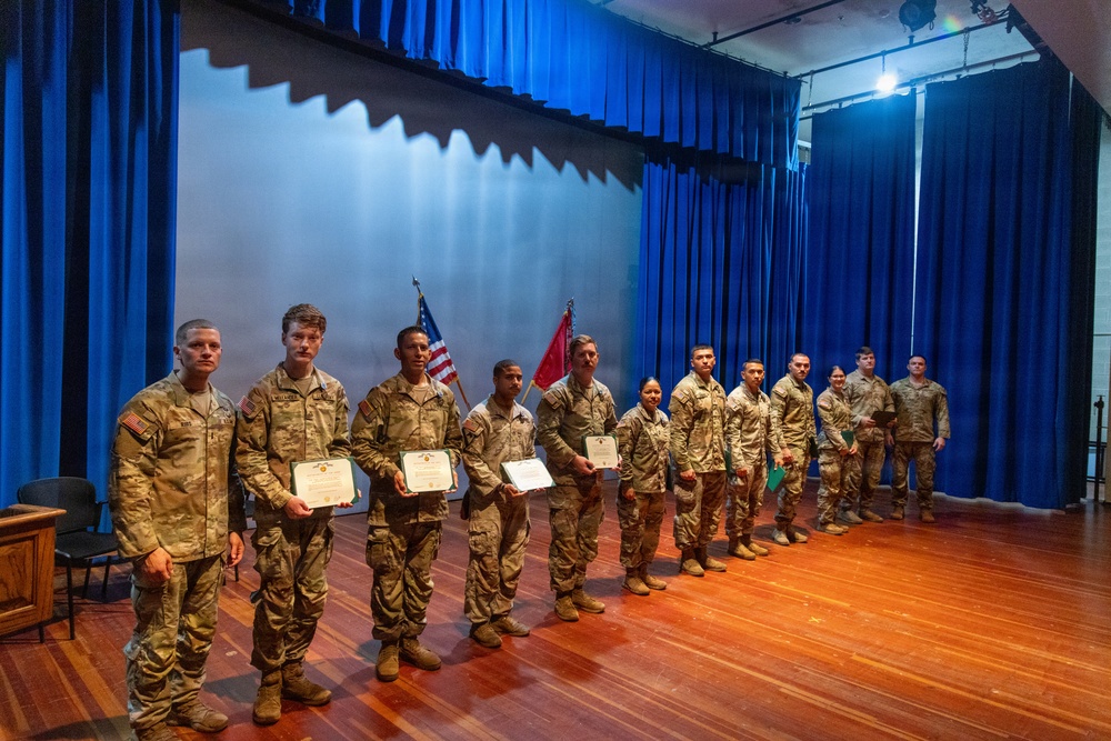 A group of military personnel in U.S. Army combat uniforms (OCP/MultiCam) are lined up on a wooden stage in front of blue curtains during what appears to be an awards or recognition ceremony. Several soldiers in the front of the line are holding certificates or commendation documents. Two flags — an American flag and what appears to be a unit or Army flag — are visible in the background.