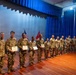 A group of military personnel in U.S. Army combat uniforms (OCP/MultiCam) are lined up on a wooden stage in front of blue curtains during what appears to be an awards or recognition ceremony. Several soldiers in the front of the line are holding certificates or commendation documents. Two flags — an American flag and what appears to be a unit or Army flag — are visible in the background.