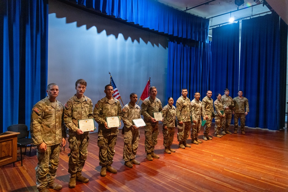 A group of approximately 12 military personnel in U.S. Army camouflage uniforms (OCP pattern) are lined up on a stage during what appears to be an awards or recognition ceremony. Several soldiers in the front are holding certificates or award documents. Two flags, including a U.S. flag and what appears to be a unit guidon, are visible in the background.