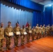 A group of approximately 12 military personnel in U.S. Army camouflage uniforms (OCP pattern) are lined up on a stage during what appears to be an awards or recognition ceremony. Several soldiers in the front are holding certificates or award documents. Two flags, including a U.S. flag and what appears to be a unit guidon, are visible in the background.