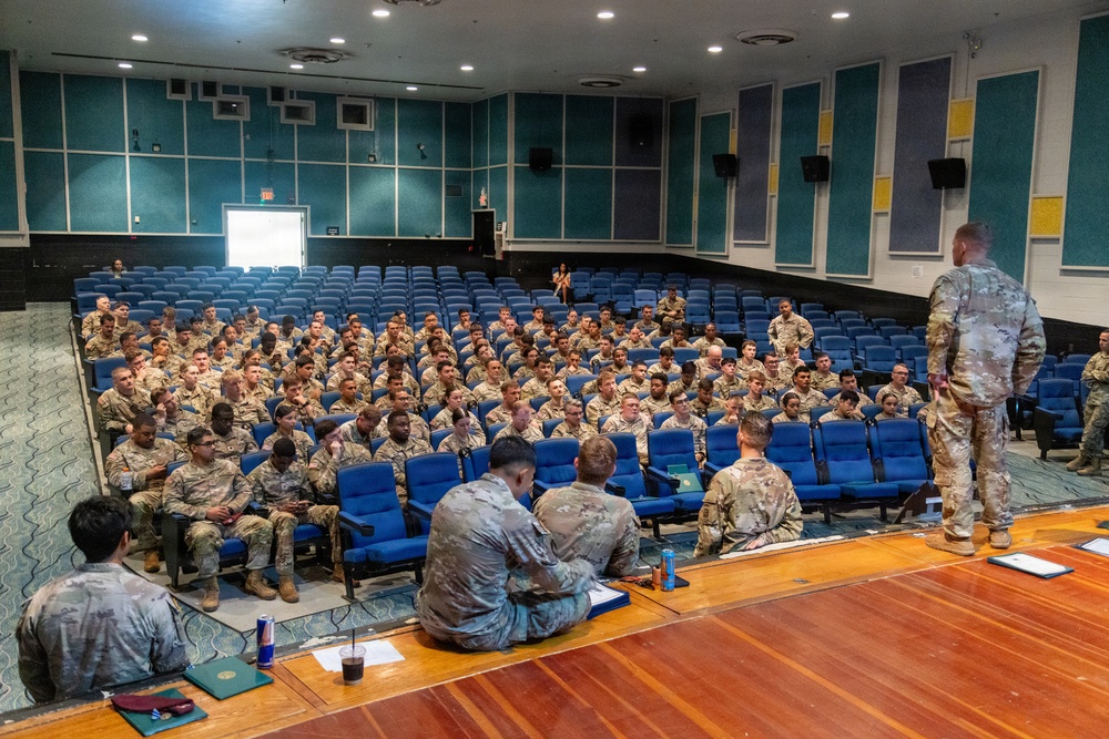 A military briefing or assembly is taking place in an auditorium with blue theater-style seating. A large group of uniformed military personnel, likely soldiers in Army Combat Uniforms (ACU/OCP pattern), are seated in the audience while one individual stands at the front addressing the group. Several personnel are seated on the stage floor facing the audience, suggesting a panel or leadership format.