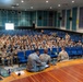A military briefing or assembly is taking place in an auditorium with blue theater-style seating. A large group of uniformed military personnel, likely soldiers in Army Combat Uniforms (ACU/OCP pattern), are seated in the audience while one individual stands at the front addressing the group. Several personnel are seated on the stage floor facing the audience, suggesting a panel or leadership format.