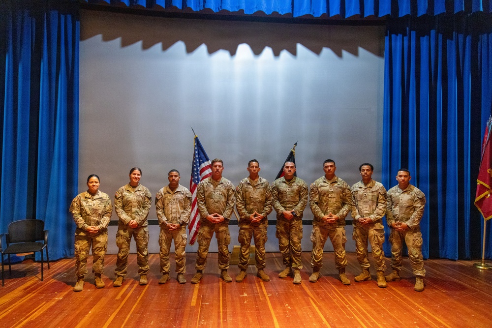 Nine military personnel in matching camouflage uniforms are posed in a formal group photograph on a stage. They are standing in a single row in front of a projection screen flanked by blue curtains, with an American flag and at least one additional flag visible behind them. The setting appears to be an auditorium or theater used for a military ceremony or event.