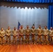 Nine military personnel in matching camouflage uniforms are posed in a formal group photograph on a stage. They are standing in a single row in front of a projection screen flanked by blue curtains, with an American flag and at least one additional flag visible behind them. The setting appears to be an auditorium or theater used for a military ceremony or event.
