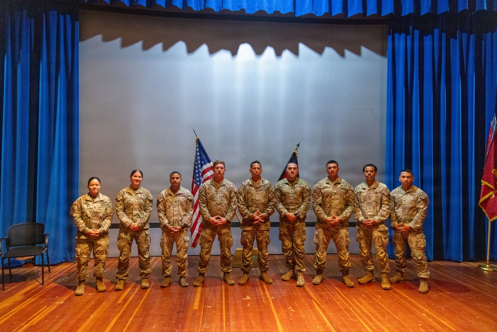 A group of nine military personnel in U.S. Army combat uniforms (OCP/MultiCam pattern) are posed for a formal group photograph on a stage. They are standing in a line in front of a projection screen flanked by blue curtains, with a U.S. flag and what appears to be a unit guidon visible behind them. The setting appears to be an auditorium or theater on a military installation.
