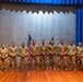A group of nine military personnel in U.S. Army combat uniforms (OCP/MultiCam pattern) are posed for a formal group photograph on a stage. They are standing in a line in front of a projection screen flanked by blue curtains, with a U.S. flag and what appears to be a unit guidon visible behind them. The setting appears to be an auditorium or theater on a military installation.
