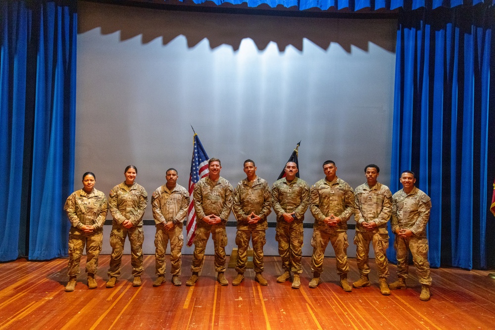 A group of ten military personnel in U.S. Army combat uniforms (OCP/MultiCam pattern) are posed together on a stage for a formal group photograph. Two flags are visible behind them — a U.S. flag and what appears to be a unit or organizational flag. The setting appears to be an auditorium or theater with blue curtains and a projection screen backdrop.