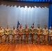 A group of ten military personnel in U.S. Army combat uniforms (OCP/MultiCam pattern) are posed together on a stage for a formal group photograph. Two flags are visible behind them — a U.S. flag and what appears to be a unit or organizational flag. The setting appears to be an auditorium or theater with blue curtains and a projection screen backdrop.