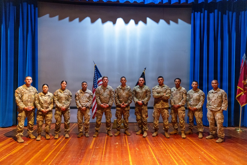 A group of eleven military personnel in U.S. Army combat uniforms (OCP/MultiCam pattern) pose for a formal group photograph on a stage. They are standing in a single row in front of a projection screen flanked by blue curtains, with a U.S. flag and what appears to be a unit guidon visible behind them. The setting appears to be an auditorium or theater on a military installation.