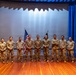 A group of eleven military personnel in U.S. Army combat uniforms (OCP/MultiCam pattern) pose for a formal group photograph on a stage. They are standing in a single row in front of a projection screen flanked by blue curtains, with a U.S. flag and what appears to be a unit guidon visible behind them. The setting appears to be an auditorium or theater on a military installation.