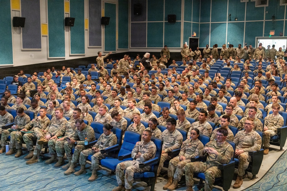A large auditorium filled with hundreds of military personnel in U.S. Army OCP (Operational Camouflage Pattern) uniforms seated in blue theater-style chairs. The gathering appears to be a formal military briefing, orientation, or training event held in a modern facility. The audience is predominantly attentive and facing toward the front of the room.