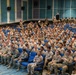 A large auditorium filled with hundreds of military personnel in U.S. Army OCP (Operational Camouflage Pattern) uniforms seated in blue theater-style chairs. The gathering appears to be a formal military briefing, orientation, or training event held in a modern facility. The audience is predominantly attentive and facing toward the front of the room.