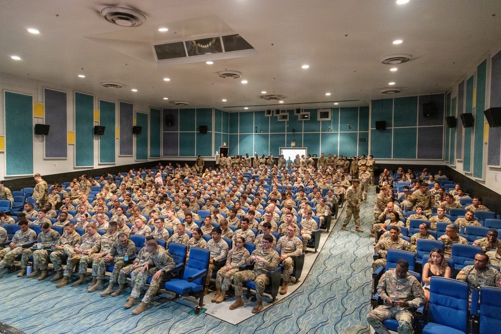 A large auditorium or theater is filled with hundreds of military personnel in camouflage uniforms seated in blue chairs. The venue appears to be a modern briefing or presentation hall with teal acoustic panels on the walls and recessed lighting. The gathering suggests a formal military briefing, orientation, or all-hands assembly.