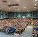 A large auditorium or theater is filled with hundreds of military personnel in camouflage uniforms seated in blue chairs. The venue appears to be a modern briefing or presentation hall with teal acoustic panels on the walls and recessed lighting. The gathering suggests a formal military briefing, orientation, or all-hands assembly.