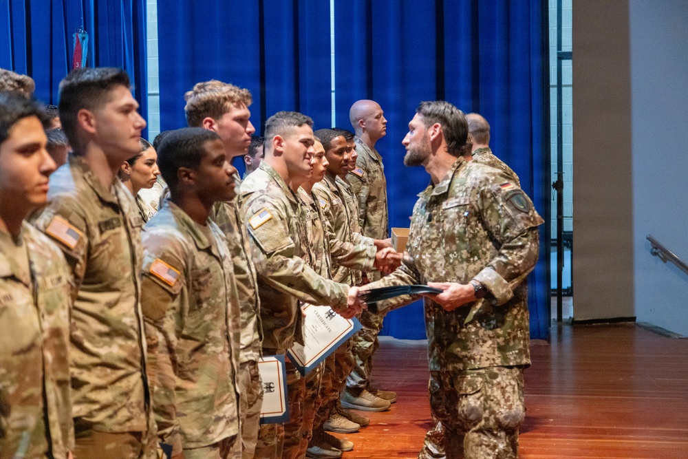A military ceremony is taking place in an indoor venue where a senior officer in what appears to be German Bundeswehr camouflage uniform is presenting certificates to a line of U.S. Army soldiers. The soldiers are standing in formation wearing U.S. Army OCP (Operational Camouflage Pattern) uniforms with American flag patches visible on their sleeves. This appears to be a graduation or award ceremony involving multinational military personnel.