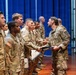 A military ceremony is taking place in an indoor venue where a senior officer in what appears to be German Bundeswehr camouflage uniform is presenting certificates to a line of U.S. Army soldiers. The soldiers are standing in formation wearing U.S. Army OCP (Operational Camouflage Pattern) uniforms with American flag patches visible on their sleeves. This appears to be a graduation or award ceremony involving multinational military personnel.