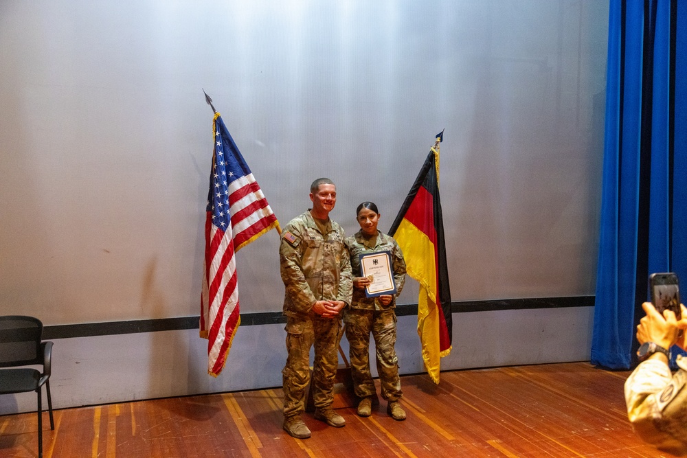 Two U.S. military personnel in camouflage uniforms are posing on a stage during what appears to be an award or recognition ceremony. One individual is holding a framed certificate or award document. The scene is flanked by a U.S. flag and a German flag, suggesting a joint U.S.-German military event.