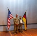 Two U.S. military personnel in camouflage uniforms are posing on a stage during what appears to be an award or recognition ceremony. One individual is holding a framed certificate or award document. The scene is flanked by a U.S. flag and a German flag, suggesting a joint U.S.-German military event.