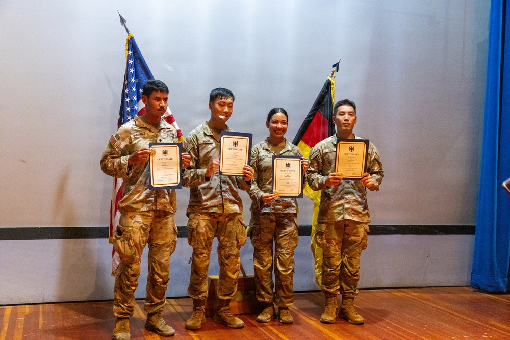 Four military personnel in camouflage uniforms are posing together on a stage, each holding a certificate. They are standing in front of an American flag and a German flag, suggesting a joint military ceremony or award presentation. All four individuals appear to be smiling, indicating a celebratory occasion.