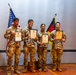 Four military personnel in camouflage uniforms are posing together on a stage, each holding a certificate. They are standing in front of an American flag and a German flag, suggesting a joint military ceremony or award presentation. All four individuals appear to be smiling, indicating a celebratory occasion.