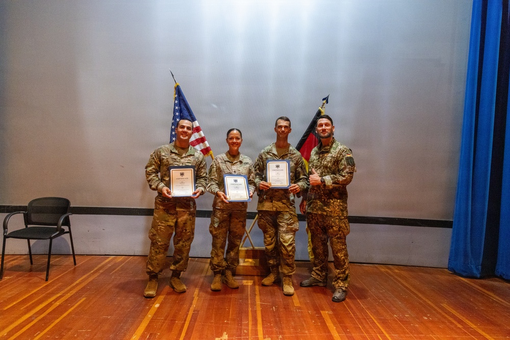 Four military personnel in camouflage uniforms are posing on a stage for a group photo. Three of them are holding framed certificates that appear to bear a German federal eagle emblem. The scene suggests a joint military ceremony involving both American and German forces, as evidenced by the US and German flags displayed behind them.