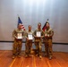 Four military personnel in camouflage uniforms are posing on a stage for a group photo. Three of them are holding framed certificates that appear to bear a German federal eagle emblem. The scene suggests a joint military ceremony involving both American and German forces, as evidenced by the US and German flags displayed behind them.