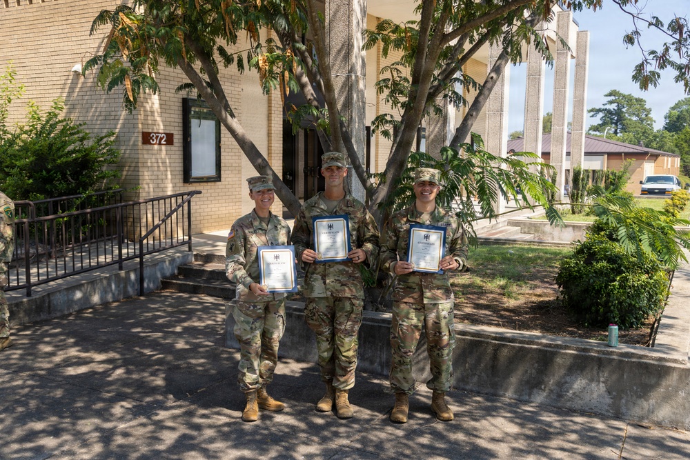 Three military personnel in U.S. Army OCP (Operational Camouflage Pattern) uniforms are posing together in front of Building 372, each holding what appears to be a certificate or award in a blue folder. The setting appears to be a military installation, likely a U.S. Army base, on a sunny day. The occasion appears to be a formal recognition or award ceremony.