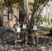 Three military personnel in U.S. Army OCP (Operational Camouflage Pattern) uniforms are posing together in front of Building 372, each holding what appears to be a certificate or award in a blue folder. The setting appears to be a military installation, likely a U.S. Army base, on a sunny day. The occasion appears to be a formal recognition or award ceremony.