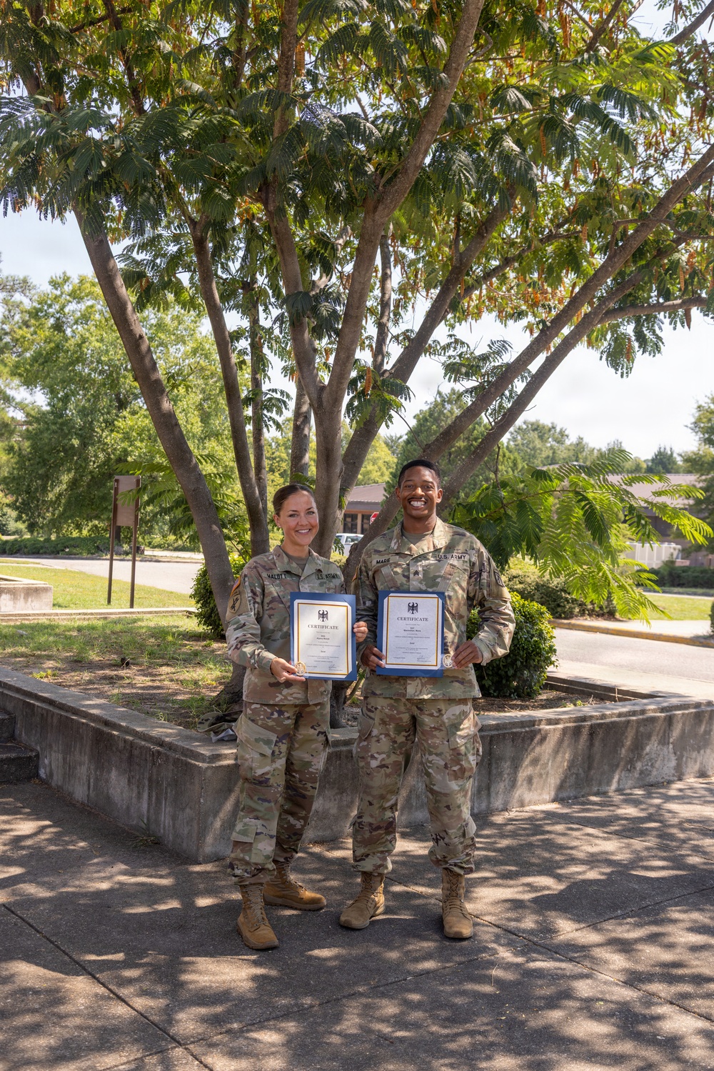 Two U.S. Army soldiers in OCP (Operational Camouflage Pattern) uniforms are posing outdoors while holding framed certificates with blue borders. Both individuals are smiling, suggesting a celebratory or recognition occasion. The setting appears to be a military installation with trees, landscaping, and paved walkways visible in the background.